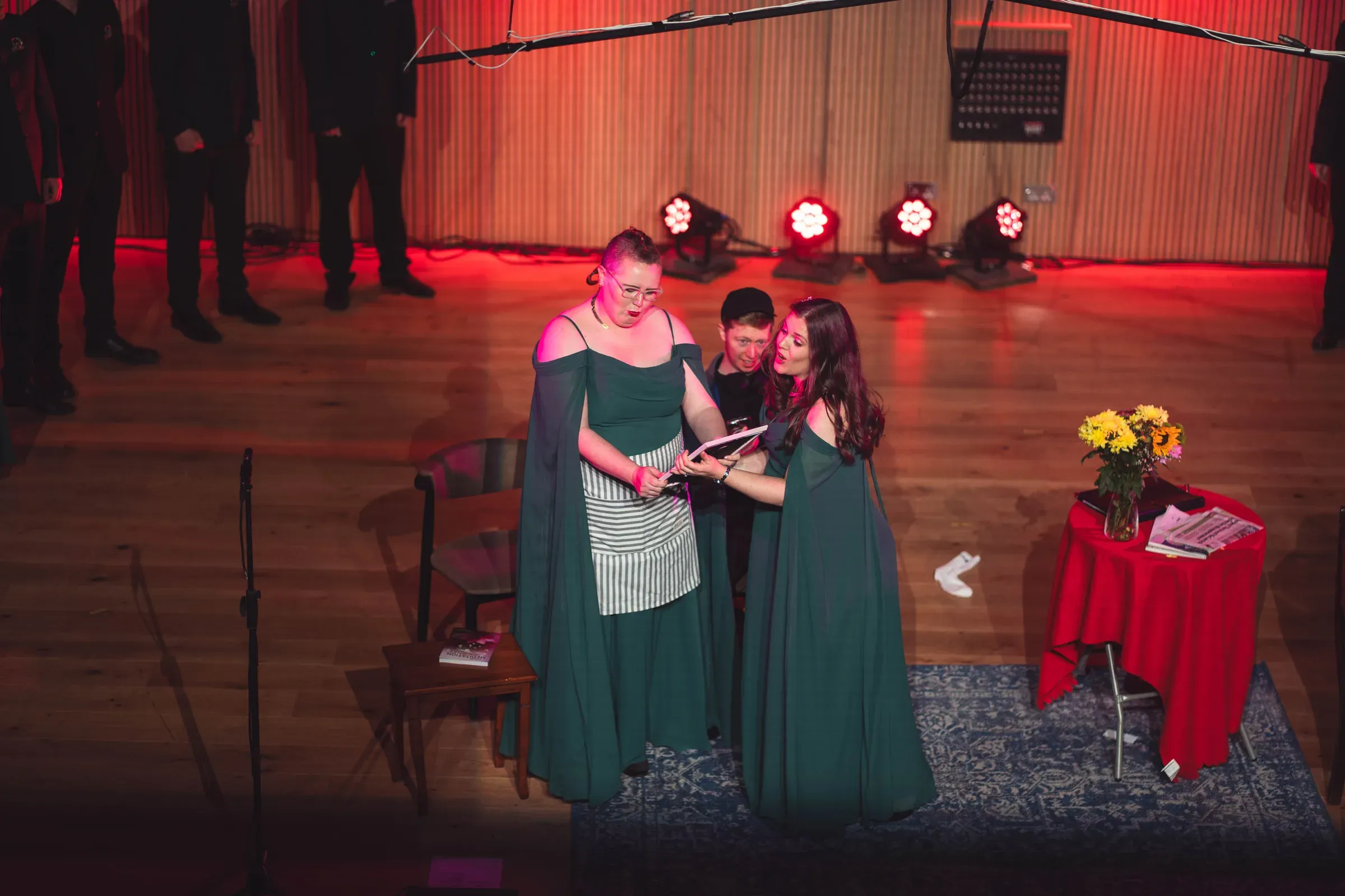 Two singers in green gowns performing a scene beside a table of flowers