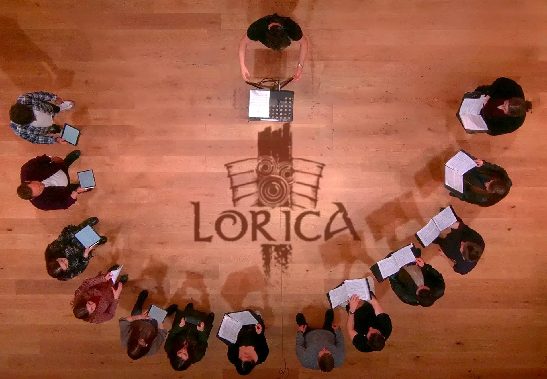 Overhead view of the choir standing in a circle around the LORICA emblem on a wooden floor